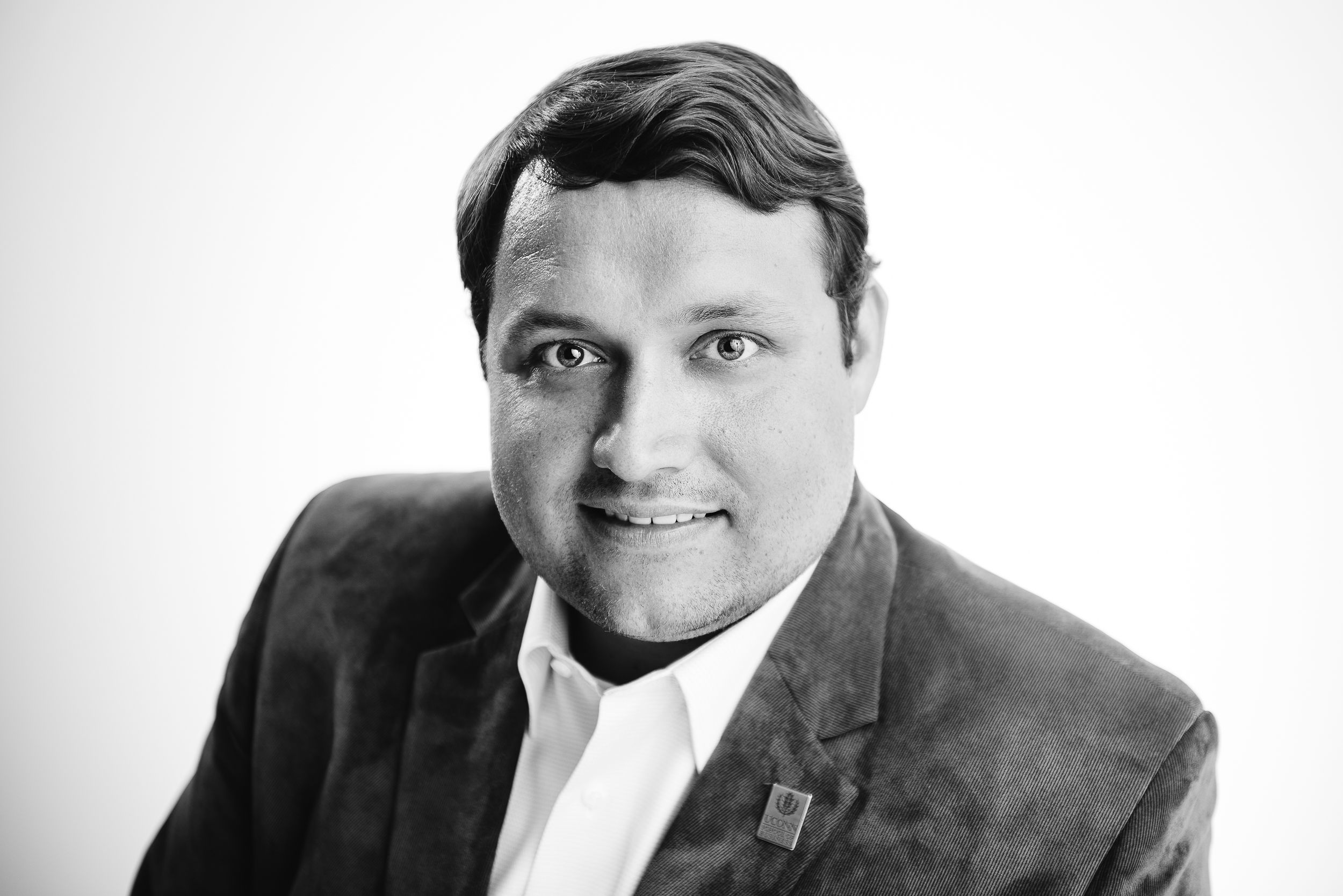 Professional black and white headshot of man dressed in professional attire with UConn pin on lapel
